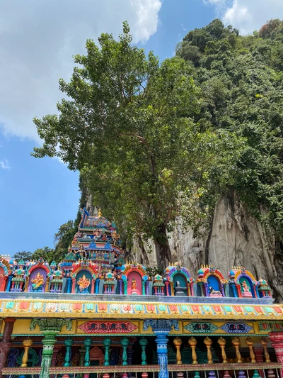 Lebendiger Hindu-Tempel, geschmückt mit komplizierten Farben, eingebettet gegen eine dramatische Kalkstein-Klippe und einen üppigen Baum unter einem strahlend blauen Himmel.