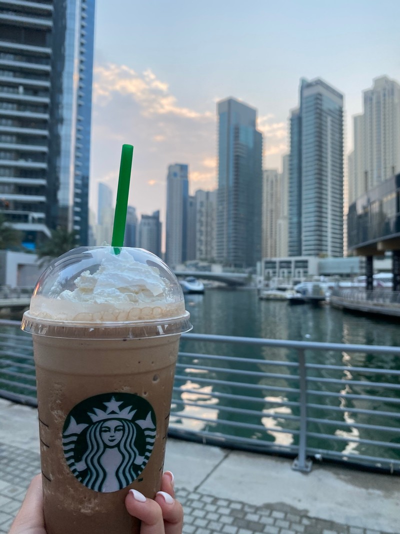 Ледяной кофе starbucks в пластиковом стакане с трубочкой на фоне городского горизонта (дубай, dubai, вода, башенный блок, напиток)