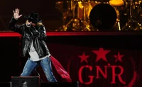 Axl Rose se apresentando apaixonadamente no palco durante um show do Guns N' Roses, com o icônico logo do GN'R e um baterista visível ao fundo.