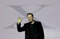 Elon Musk gestures while speaking at a Tesla event, showcasing his formal attire against a backdrop featuring the Tesla logo.