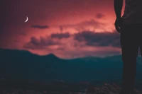 A silhouetted figure stands against a vibrant red sky at dusk, where a crescent moon hangs above the horizon, evoking a serene atmosphere.