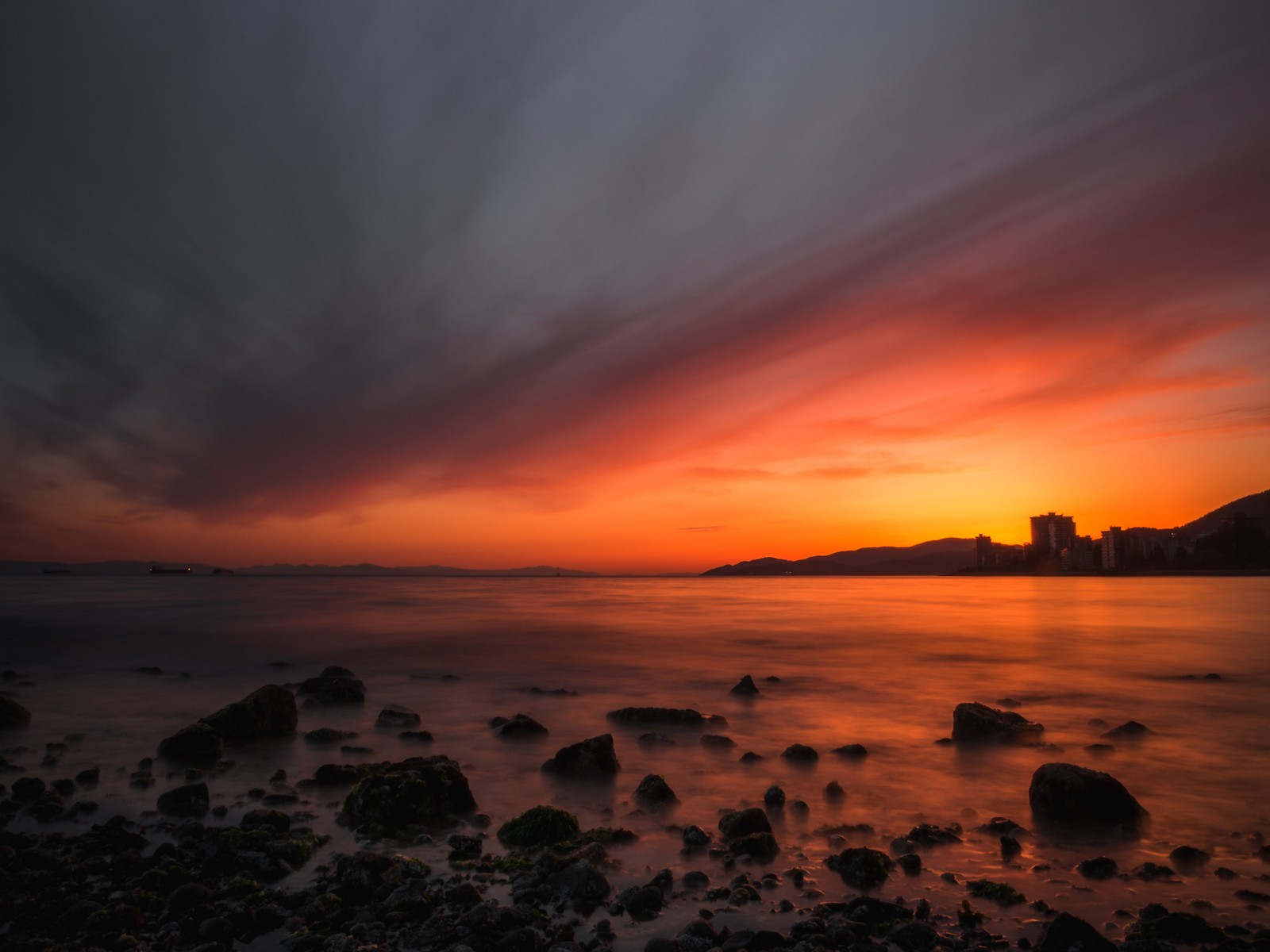 Uma vista de um pôr do sol sobre o oceano com pedras e uma cidade ao longe (por do sol, mar, horizonte, costa, resplendor)