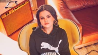 Selena Gomez en una elegante sudadera de Puma, sentada en un acogedor ambiente inspirado en lo retro.