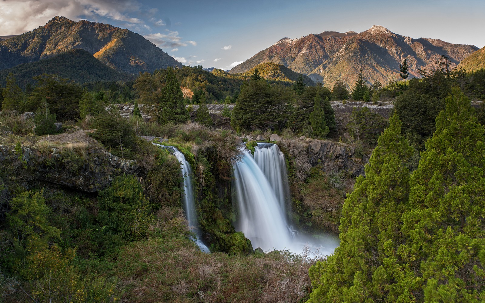 Una cascada en medio de un bosque con montañas al fondo (naturaleza, cascada, cuerpo de agua, montaña, agua)