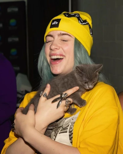 Billie Eilish lächelt mit einem grauen Kätzchen in ihren Armen