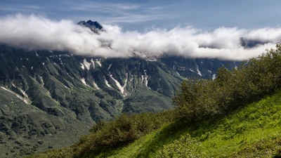 Lush green slopes meet dramatic volcanic ridges under a cloudy sky in the Kamchatka Peninsula.