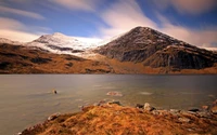 Snow-capped mountains rise majestically above a tranquil loch, reflecting the serene beauty of the highland wilderness under a dynamic sky.