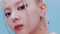 Lia from ITZY showcases a striking close-up with a bold makeup look and unique hairstyle against a soft blue background.