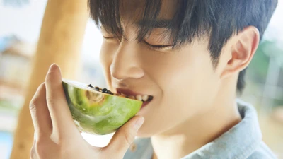 Dawon of SF9 enjoying a slice of watermelon, smiling warmly in a bright, sunny setting.