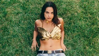 Camila Mendes in a golden shell top and denim shorts, lying on lush green grass.