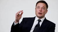 Elon Musk speaking in a suit, emphasizing his points during a business event.