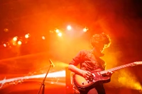 Guitarrista dinâmico se apresentando energeticamente no palco em meio a luzes vibrantes e fumaça em um concerto de rock.