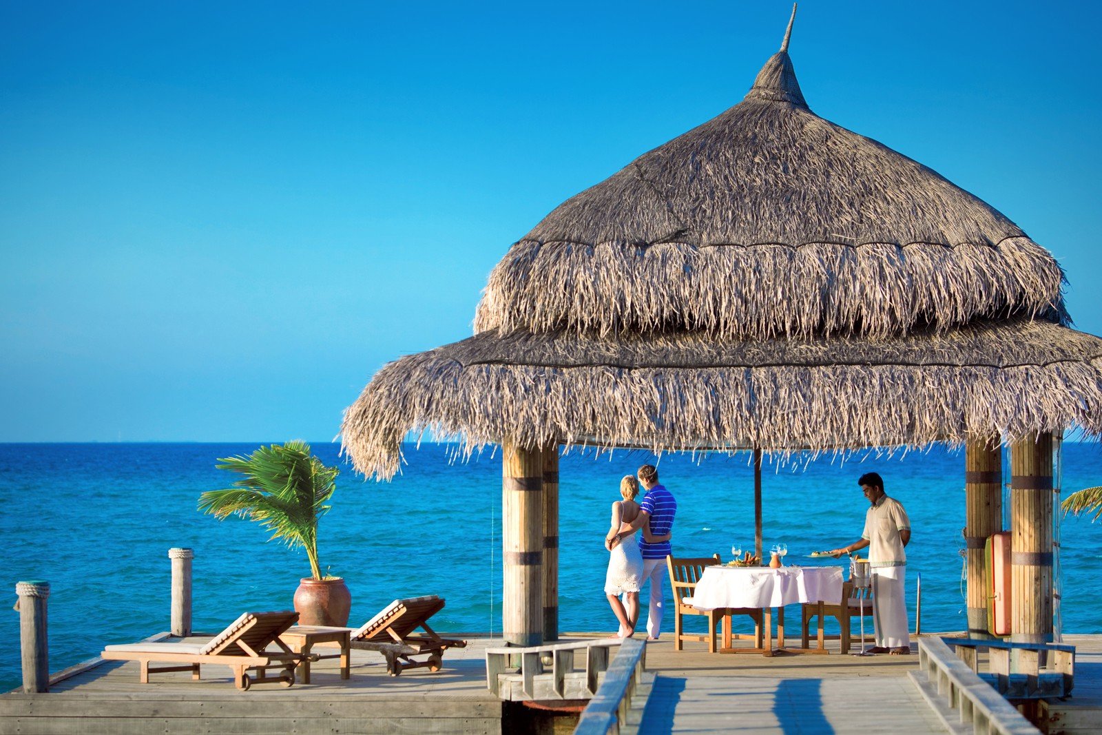 Hay dos personas sentadas a una mesa en un muelle (atolón ari, ari atoll, complejo turístico, isla, playa)