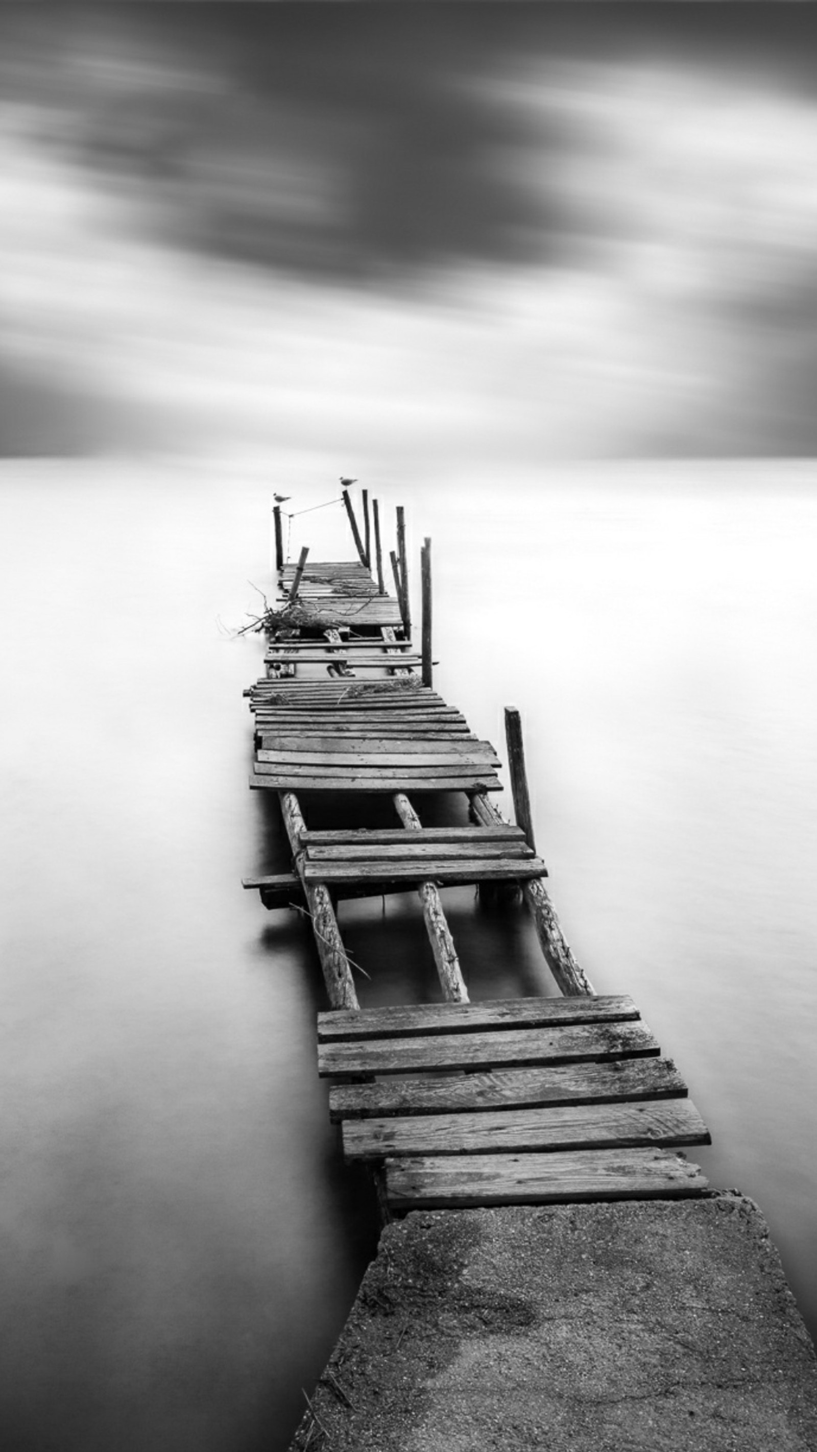 Una foto en blanco y negro de un largo muelle en el agua (back and white, roto, nubes, gris, camino)