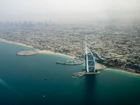 Luftaufnahme des Burj Al Arab Jumeirah mit Blick auf die Küste, mit dem Burj Khalifa in der fernen Skyline und einer ruhigen Bucht darunter.