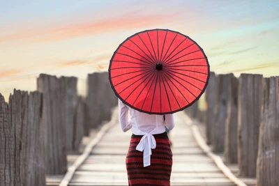 Burmesische Frau in traditioneller Kleidung, die einen roten Regenschirm auf einem Holzsteg bei Sonnenuntergang hält.