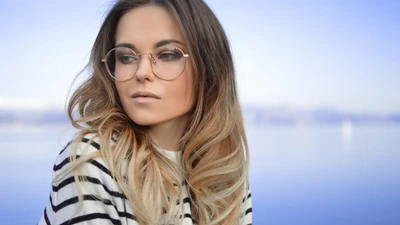 Modelo de moda elegante con cabello largo y ondulado castaño, usando gafas elegantes y una blusa a rayas, contra un fondo sereno.