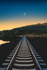 Trilhos de trem levando a um pôr do sol sereno com uma lua crescente sobre as montanhas