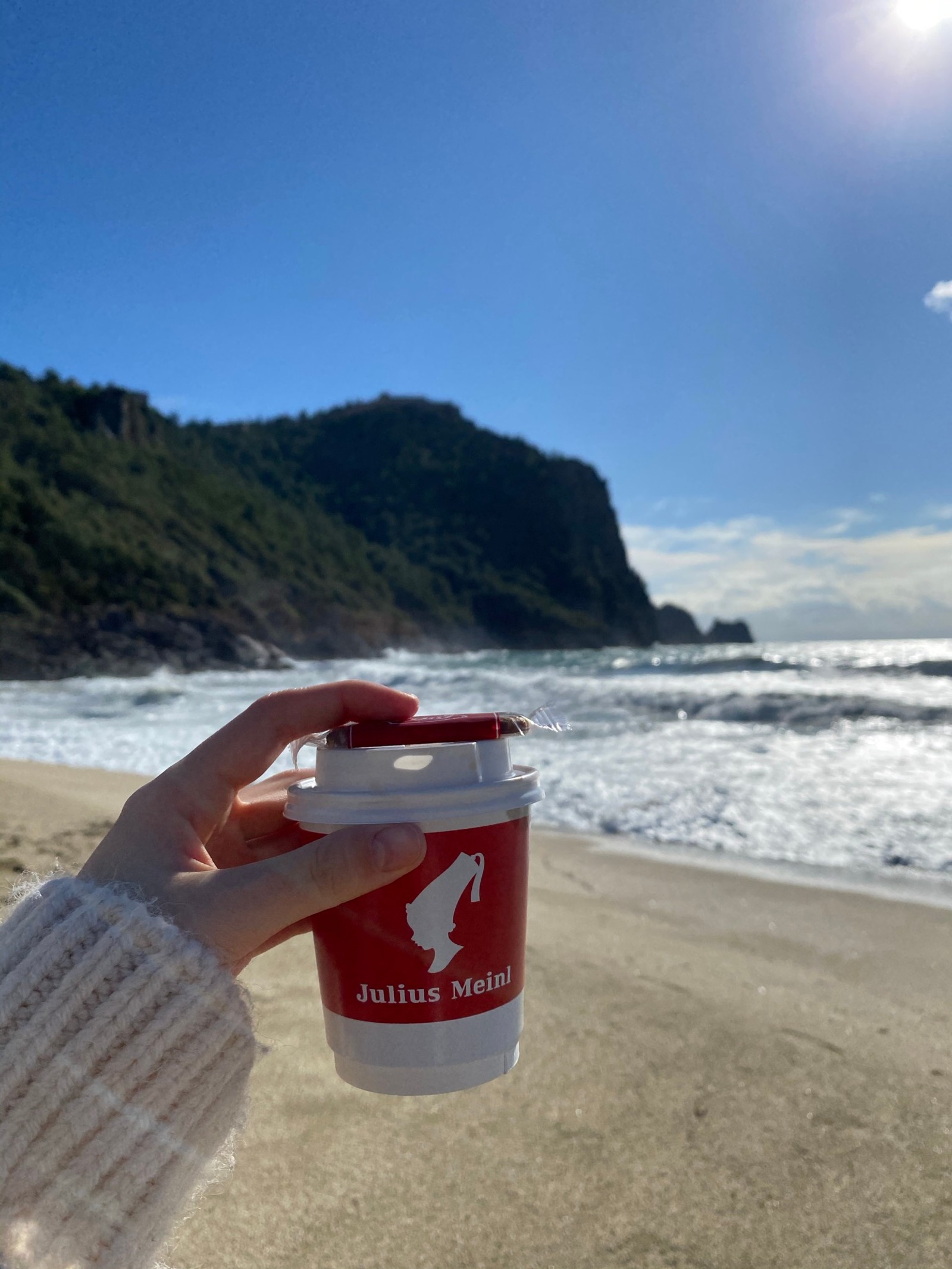 Alguien sosteniendo una taza de café en la playa con una montaña de fondo (playa, agua, nube, montaña, fluido)