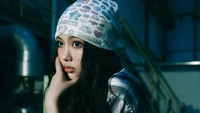 Hyein from NewJeans in a contemplative pose, wearing a patterned beanie and a plaid shirt, set against an industrial backdrop.