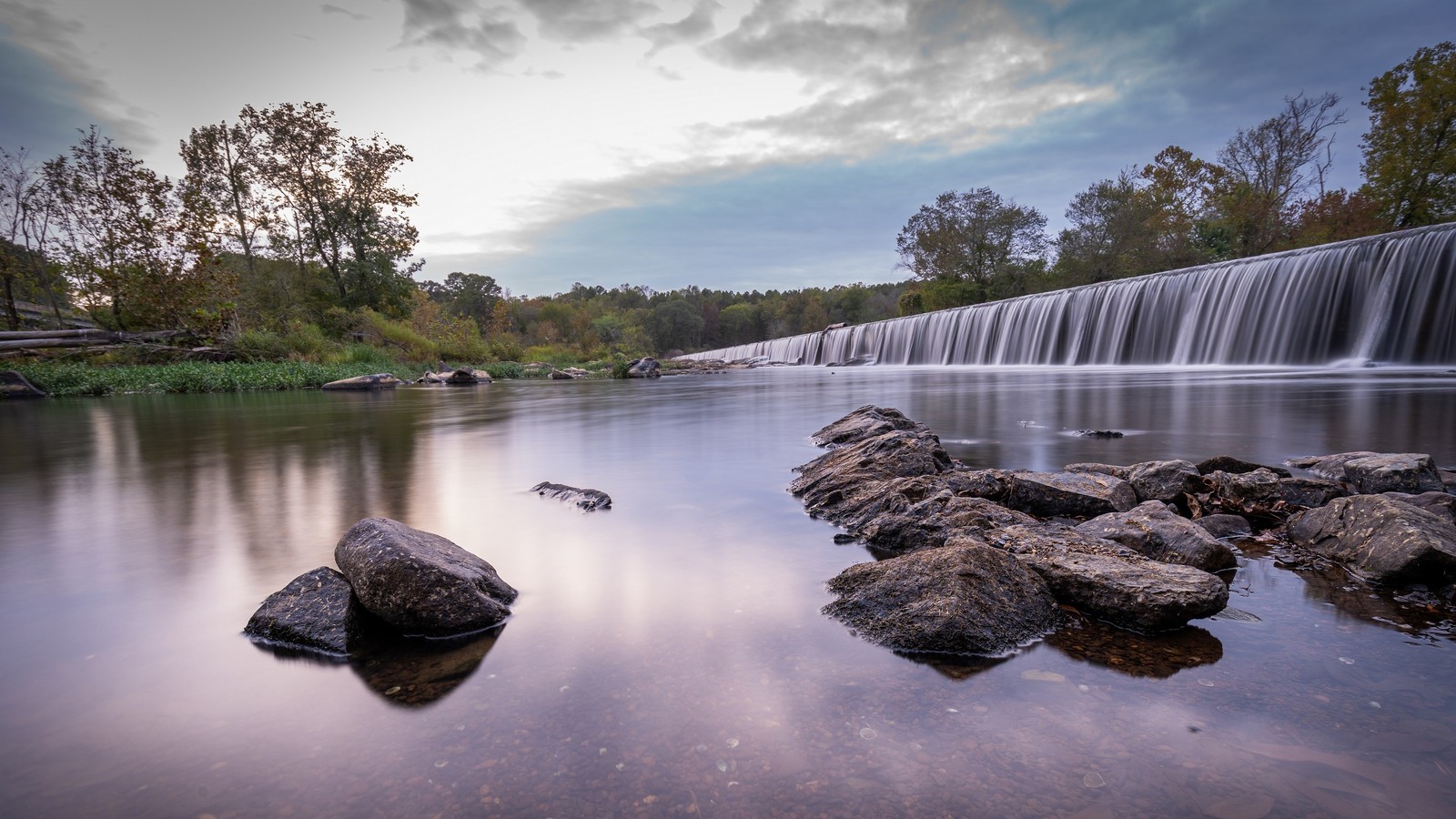 Eine sicht auf einen wasserfall mit steinen im vordergrund (reflexion, wasser, wolke, natürliche landschaft, baum)