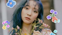 Binnie do Oh My Girl com cabelo azul, segurando flores silvestres, cercada por decorações florais coloridas.