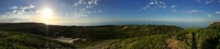 Vista panorâmica de uma paisagem costeira com vegetação exuberante, terras de pastagem e um horizonte marinho sereno sob um céu claro.