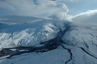 Schneebedeckter Stratovulkan mit Gletscherlandschaften und einer Wolke aus Vulkanasche, die in den bewölkten Himmel aufsteigt.