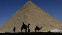 Silhuetas de camelos e turistas em frente à Grande Pirâmide de Gizé contra um céu claro, mostrando um monumento histórico da arquitetura egípcia antiga.