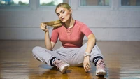 Meg Donnelly em uma pose estilosa e relaxada, vestindo uma blusa rosa e calças de moletom cinzas, exalando confiança e charme.