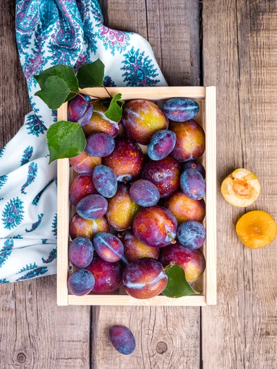 Fresh, vibrant plums nestled in a wooden crate, accompanied by a halved apricot and a decorative fabric backdrop.