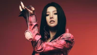 Soyeon of (G)I-DLE exudes confidence in a striking pink ensemble, holding a grenade-shaped accessory against a bold, gradient backdrop, embodying the essence of "I Never Die.