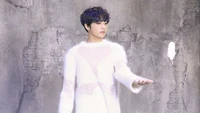 Jungkook en un suéter blanco caprichoso, elegantemente posado mientras interactúa con una pluma flotante contra un fondo texturizado.