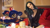 Emma Dumont en un llamativo atuendo de encaje negro, posando elegantemente en un entorno vibrante con decoración tradicional y un disfraz de danza del león en el fondo.