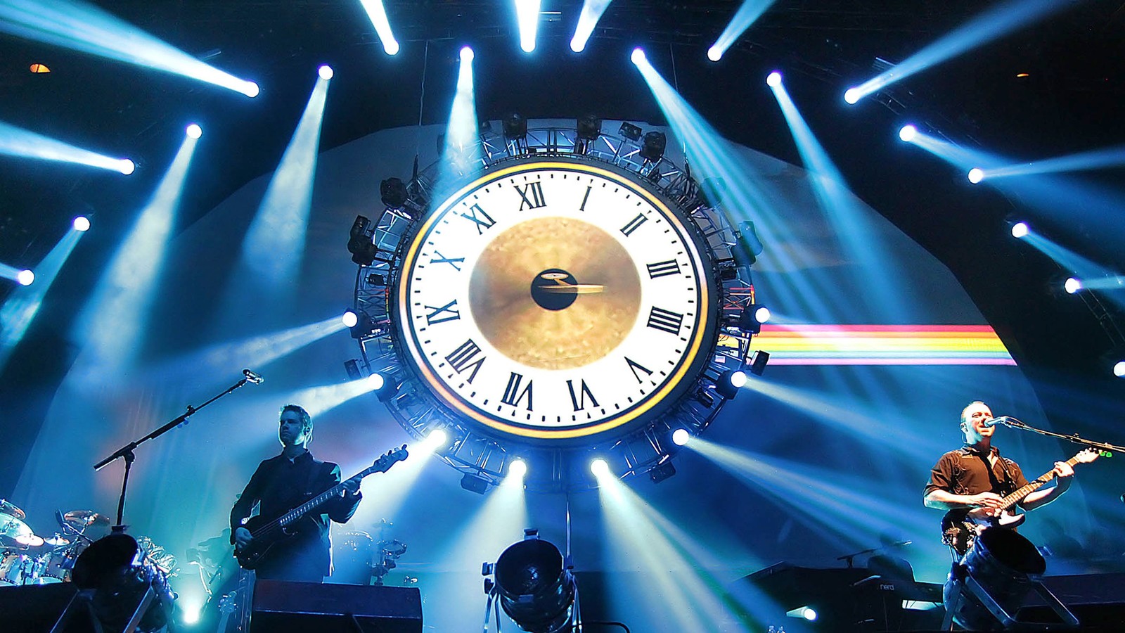 Uma imagem de um grupo de pessoas tocando no palco com um grande relógio ao fundo (pink floyd, concerto, ato de tributo, desempenho, palco)