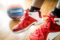 Tênis de basquete Nike em vermelho vibrante e branco, combinados com uma meia preta, pairam sobre uma bola de basquete Spalding em um piso de madeira.