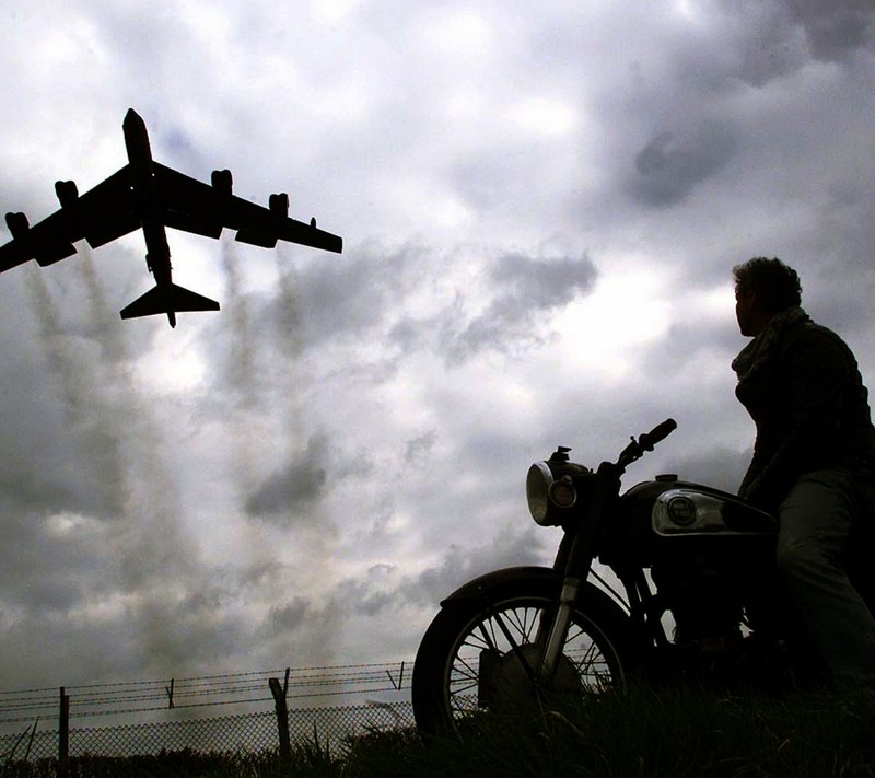 There is a man sitting on a motorcycle watching a plane fly overhead (air, bike, man, photo, plane)