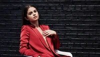 Selena Gomez in a striking red suit, exuding confidence against a dark brick backdrop.