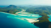 Aerial view of the Whitsunday Islands showcasing the stunning Whitehaven Beach, with its winding inlets and vibrant turquoise waters contrasting against lush green highlands and the expansive Great Barrier Reef.