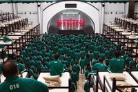 Los participantes en uniformes verdes se reúnen en un dormitorio abarrotado para un evento dramático en un programa de entretenimiento surcoreano.