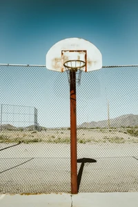 Quadra de basquete abandonada com uma cesta desgastada e cerca de arame, emoldurada por um céu limpo e montanhas distantes.