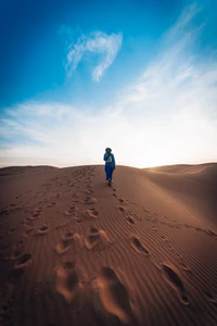 Une figure solitaire en bleu marche à travers les dunes de sable ondulantes du Sahara, sous un vaste ciel bleu orné de nuages légers.