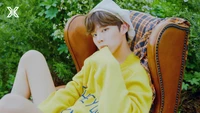 Wooseok em um suéter amarelo, relaxando em uma poltrona vintage em meio a uma vegetação exuberante.