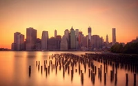 Manhattan skyline at sunrise, featuring skyscrapers reflected in the water and remnants of old piers in the foreground.