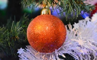 Glittering orange Christmas ornament hanging from an artificial tree, surrounded by festive decor.