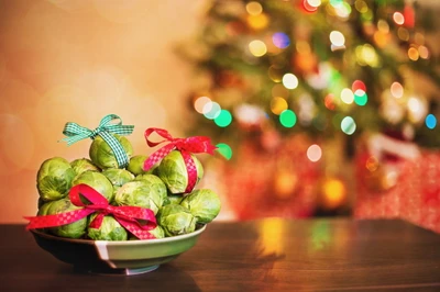 Un tazón festivo de coles de Bruselas adornado con cintas coloridas, frente a un árbol de Navidad suavemente iluminado y decoraciones navideñas.