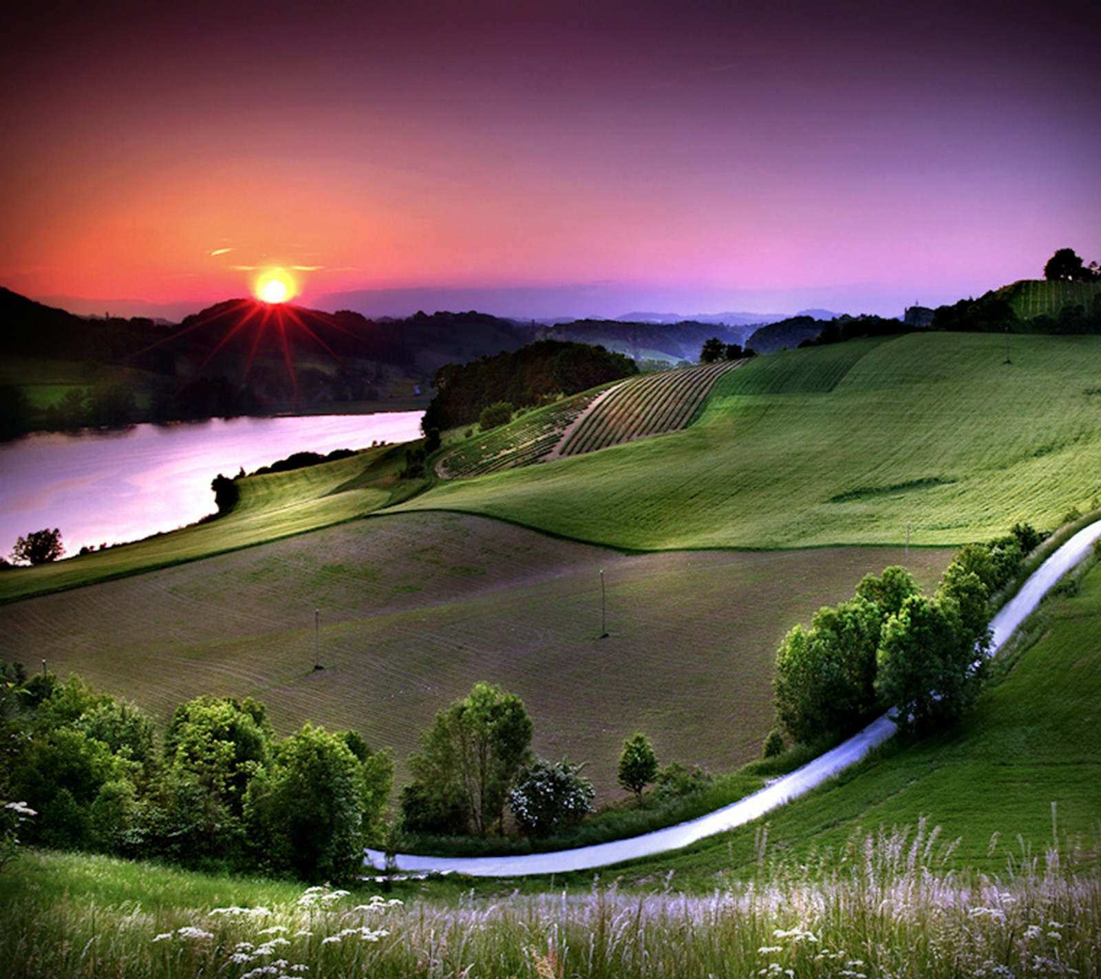 Arafed view of a winding road in a green field with a lake in the distance (breathtaking, paradise, spring, sunset)