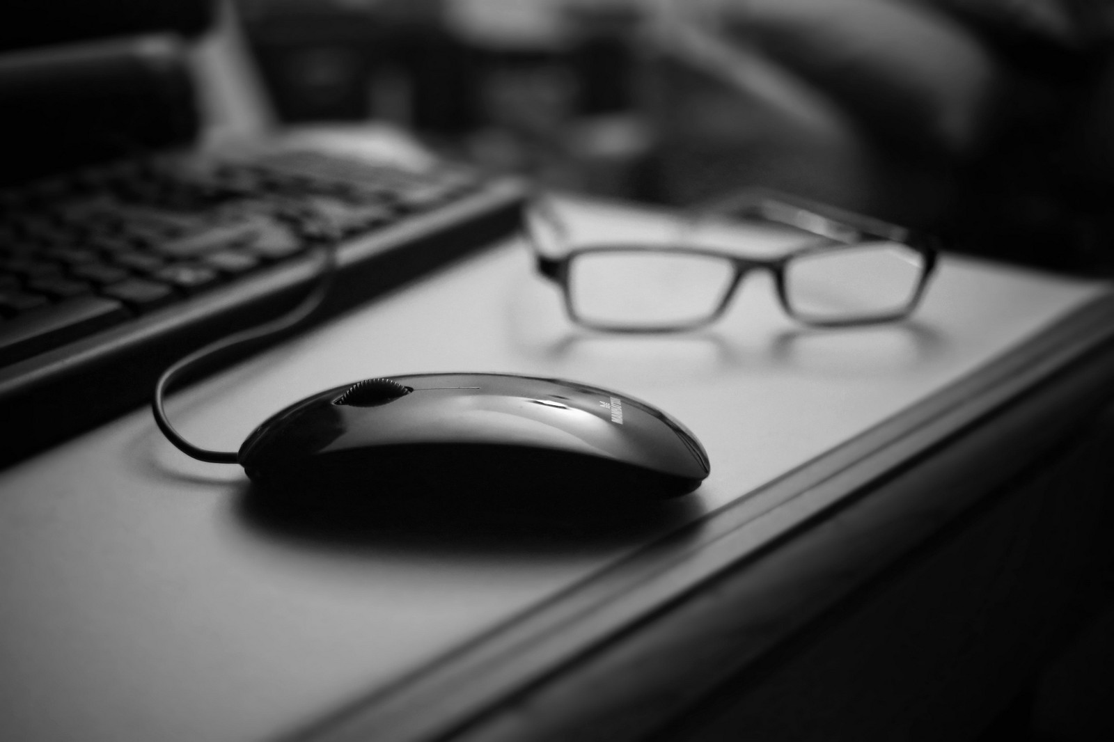 Hay un ratón y unas gafas en un escritorio junto a un teclado (negro, gafas, dispositivo electrónico, tecnología, blanco y negro)