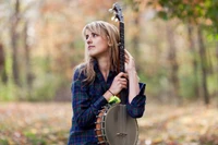 A girl with long hair holds a banjo, gazing thoughtfully amid a beautiful autumn landscape filled with colorful foliage and grasses.