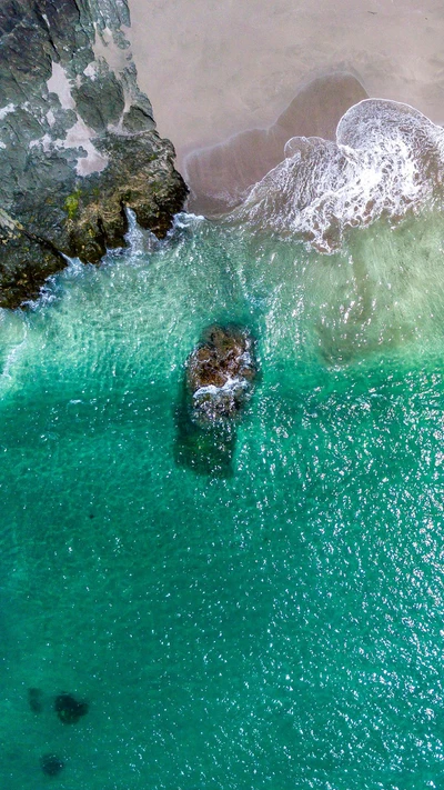 Vista aérea de uma praia tranquila com águas turquesa claras que se quebram suavemente contra formações rochosas.